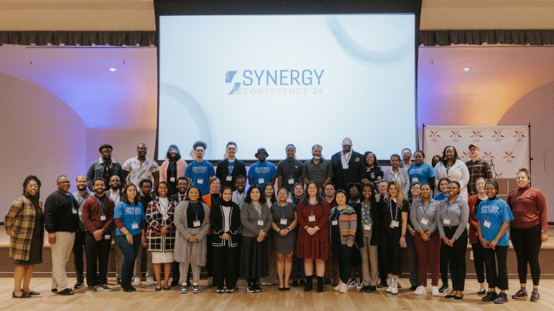 The attendees, speakers, and volunteers at the Synergy Conference gathered for a group photos in front of a screen with the conference logo
