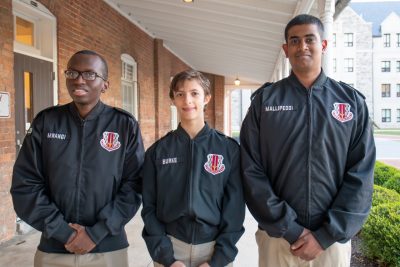 Cadets in uniform stand smiling on the covered porch of an old brick building. 