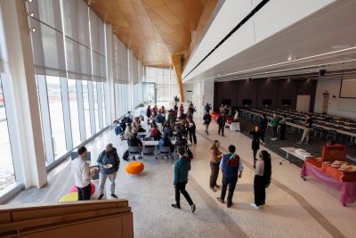 Graduate students chat and eat pizza in the atrium of the new Innovation Campus Academic Building One in Alexandria on the first day of the semester. The Boeing Auditorium is on the right and tall glass windows are on the left.