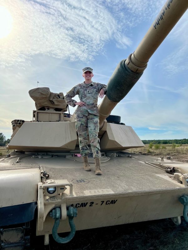 Hoffman stands atop a tank while smiling in her camouflage uniform.