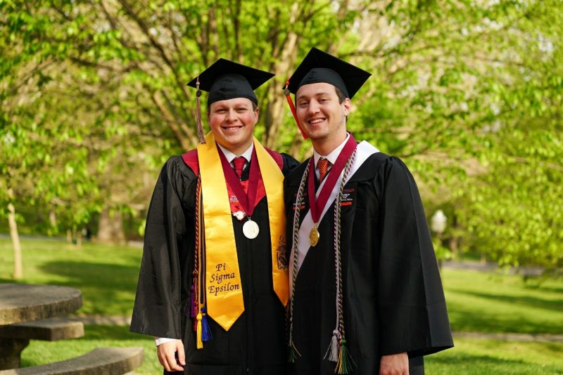Blake Warner (at left) and William Poland (at right) stand outside Hillcrest Hall in graduation regalia.