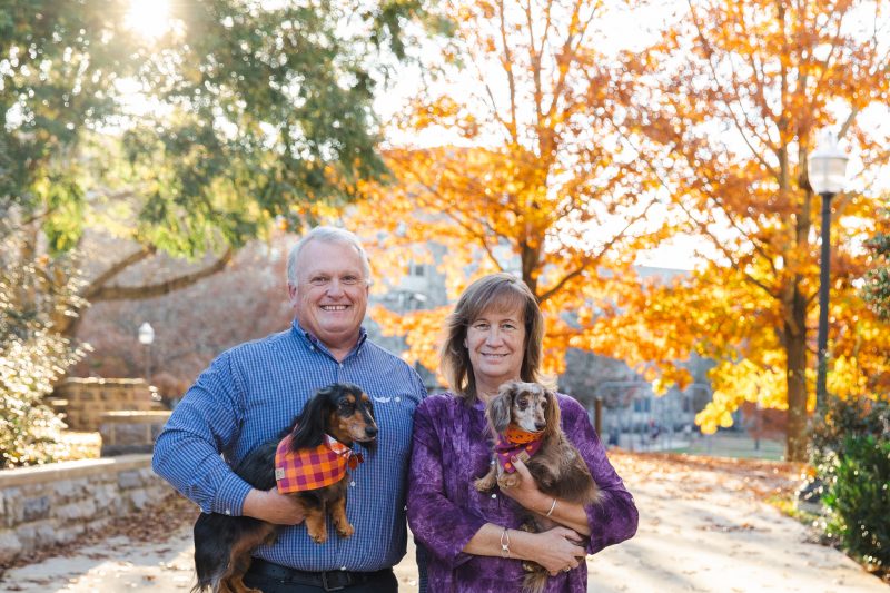 Rick Rudd and Donna Westfall-Rudd with dachshunds Annie and Buck in a backdrop of autumn sun and golden leaves.