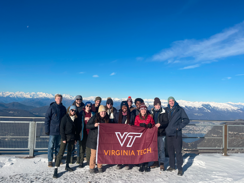 A group of students hold the Virginia Tech flag at a lookout point. A mountain range can be seen behind them.