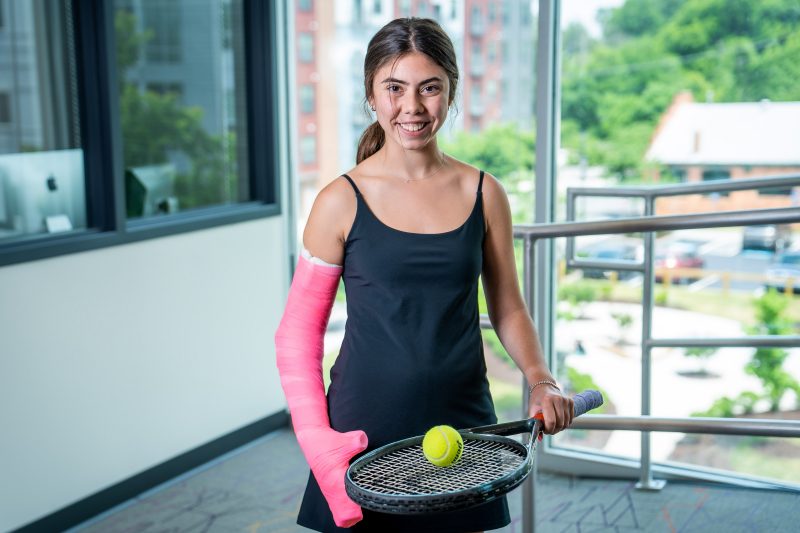 Keya Shapiro smiling while holding tennis racket and ball with pink cast on right arm.