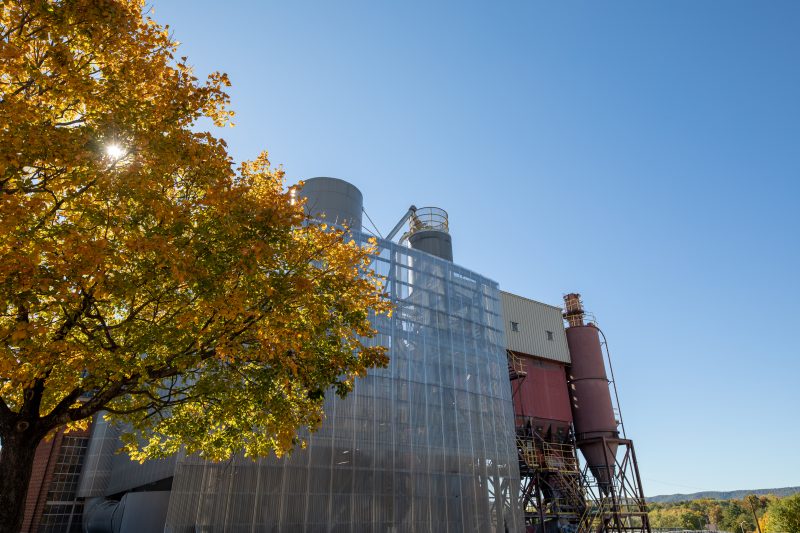 Red and grey Virginia Tech Power Plant on a sunny autumnal day. The sun shines through a tree with yellow leaves in the foreground and in the background are mountains with many trees with leaves beginning to turn yellow.
